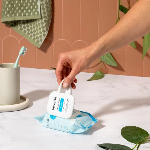 Hand holding a bottle of Honey Pot wipes on a table with a cup and toothbrush in the background.